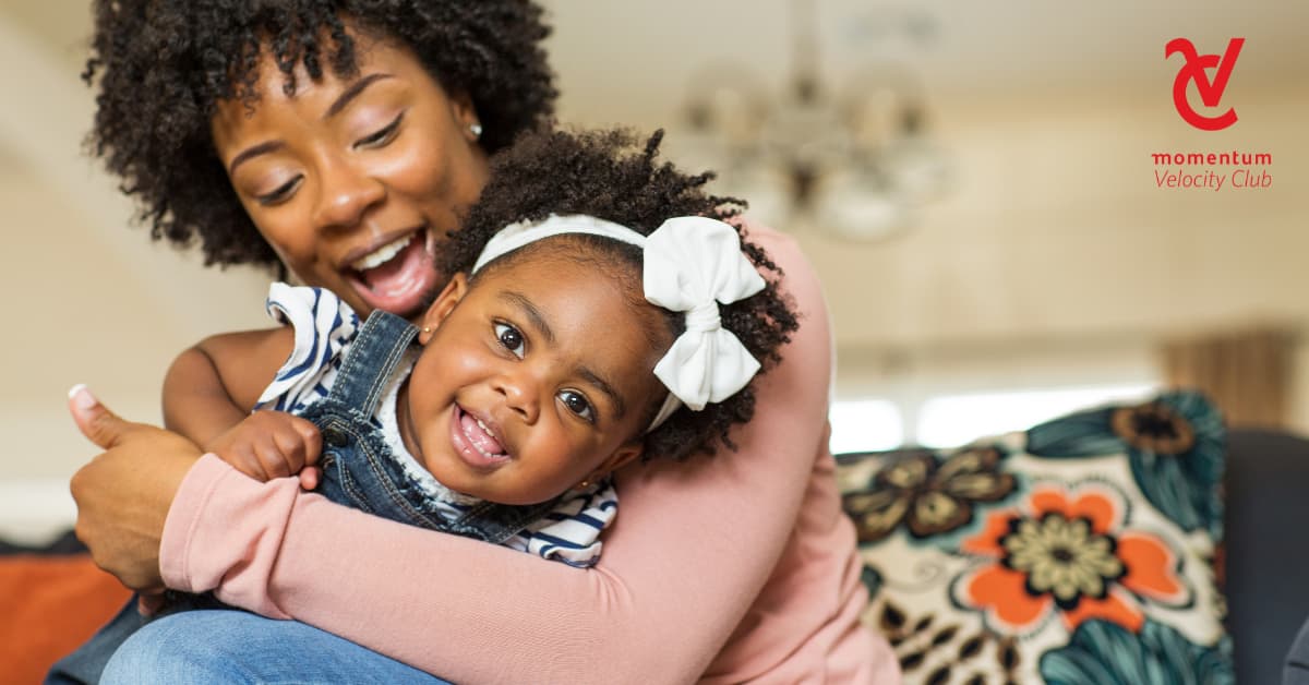 A mom is seated on a couch. She&rsquo;s holding her young daughter, and they&rsquo;re playing and having fun together.