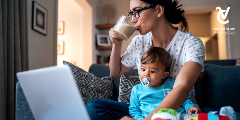A young parent is sitting on a couch and working on her budget on her laptop. She&rsquo;s drinking coffee while her young son sits next to her.